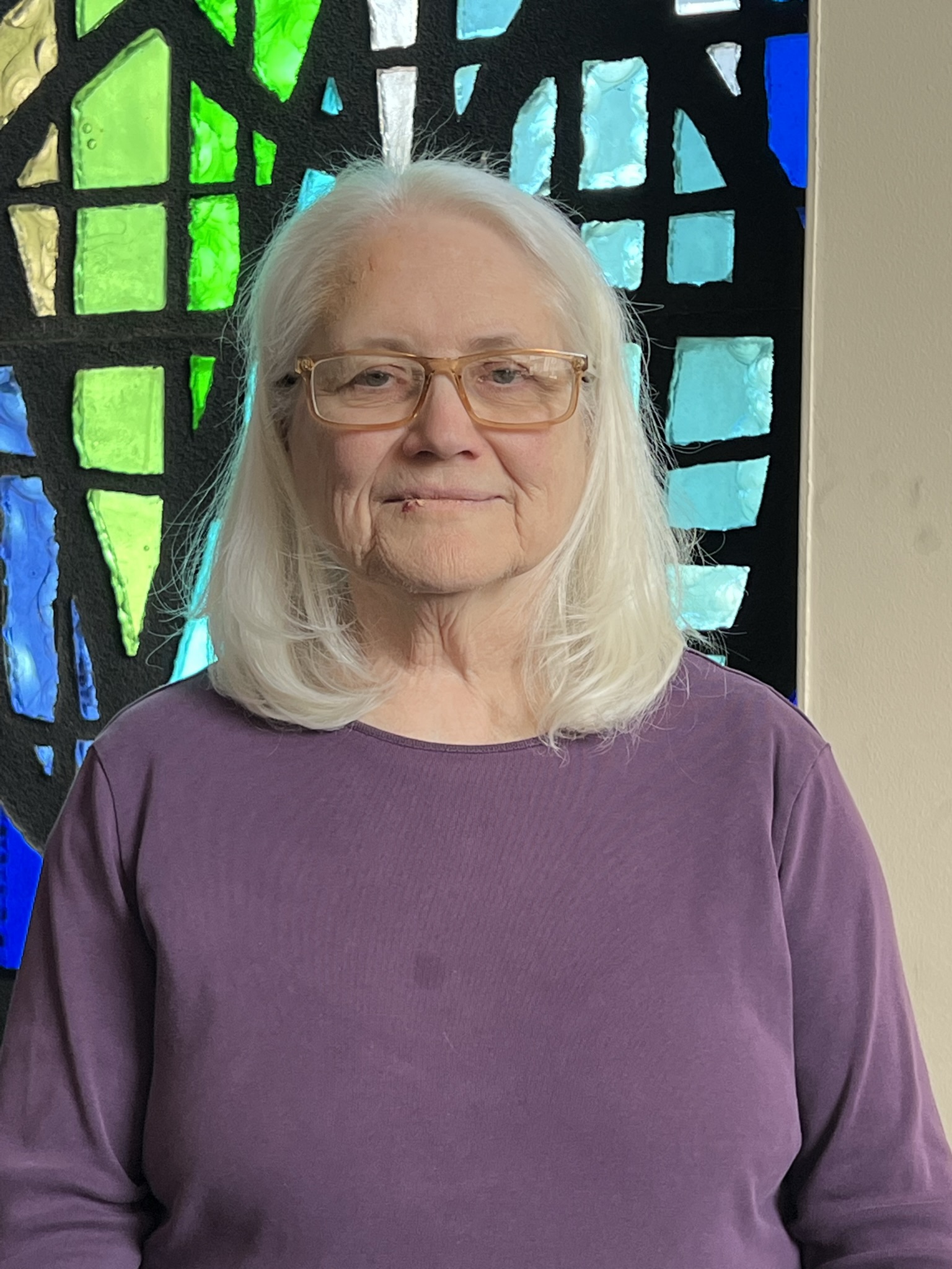 Smiling elderly woman in purple sweater in front of stained glass window.