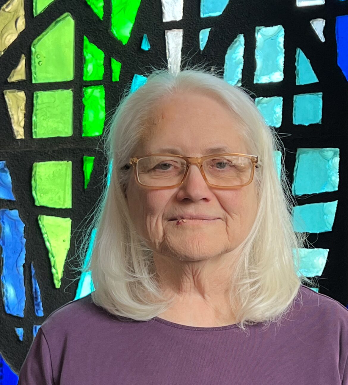 Elderly woman with white hair and glasses in front of colorful stained glass.