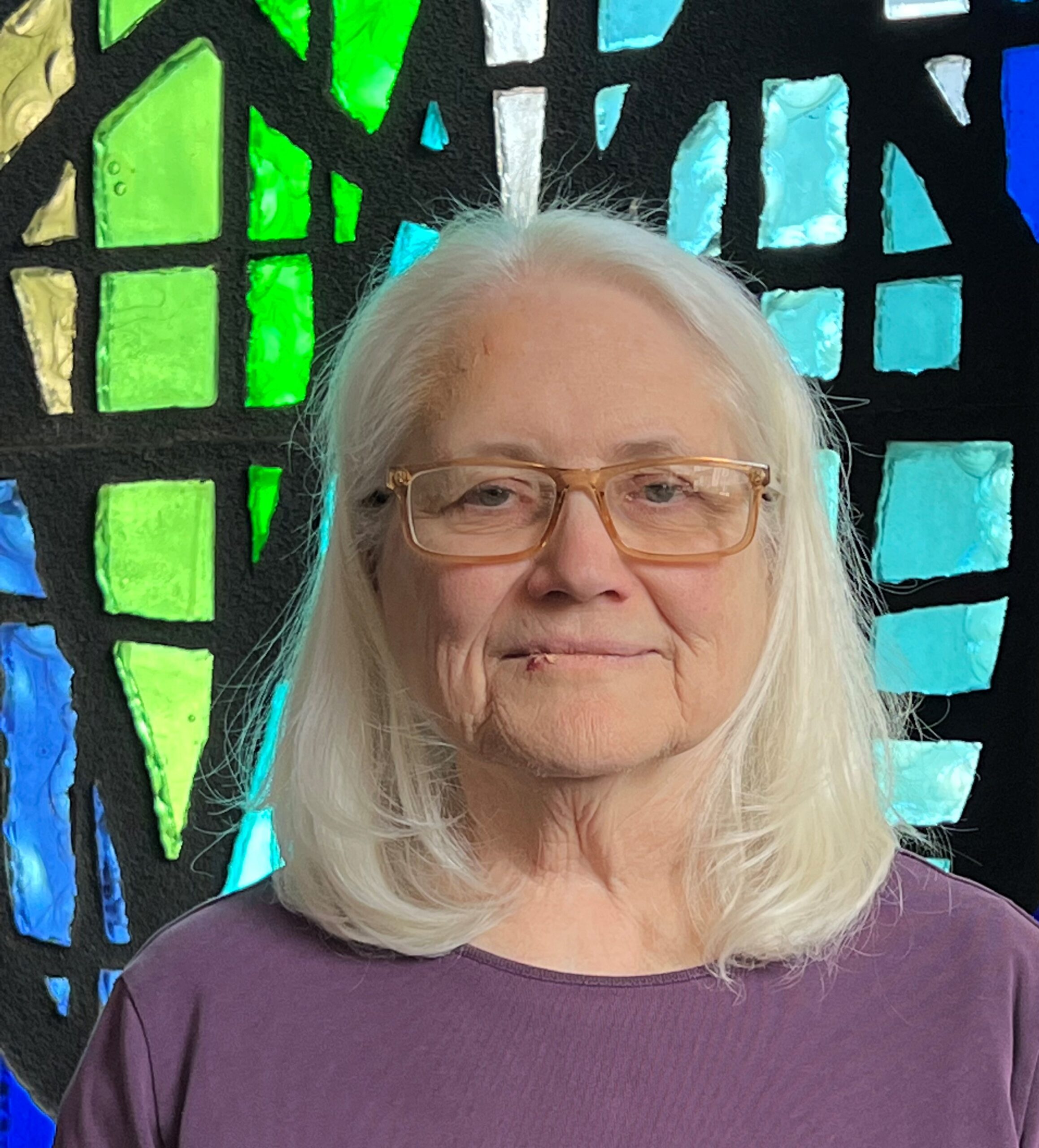 Elderly woman with white hair and glasses in front of colorful stained glass.