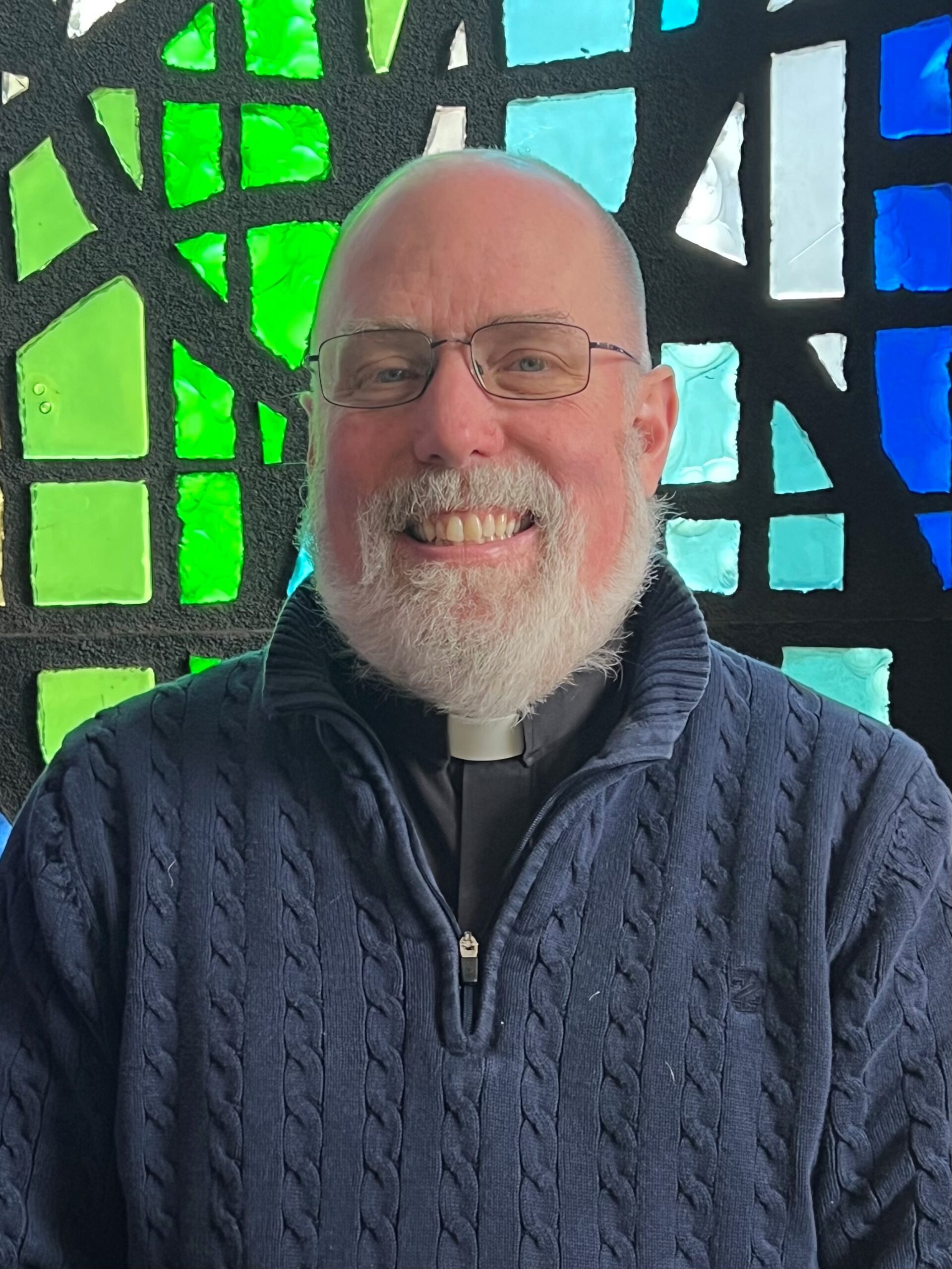 Smiling elderly man with glasses in front of colorful stained glass.