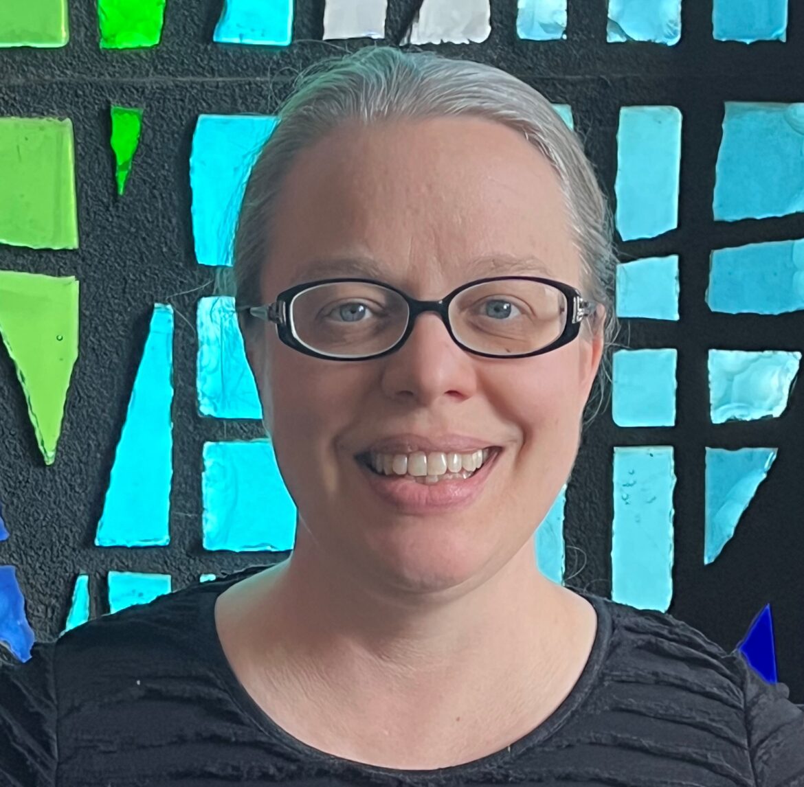 Smiling woman with glasses in front of a colorful stained-glass window.