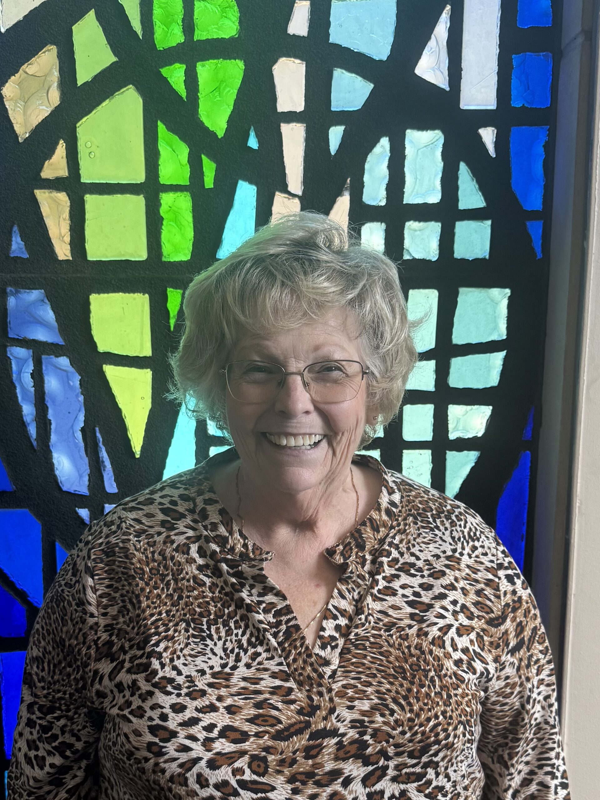 Smiling elderly woman in leopard print shirt in front of colorful stained glass.